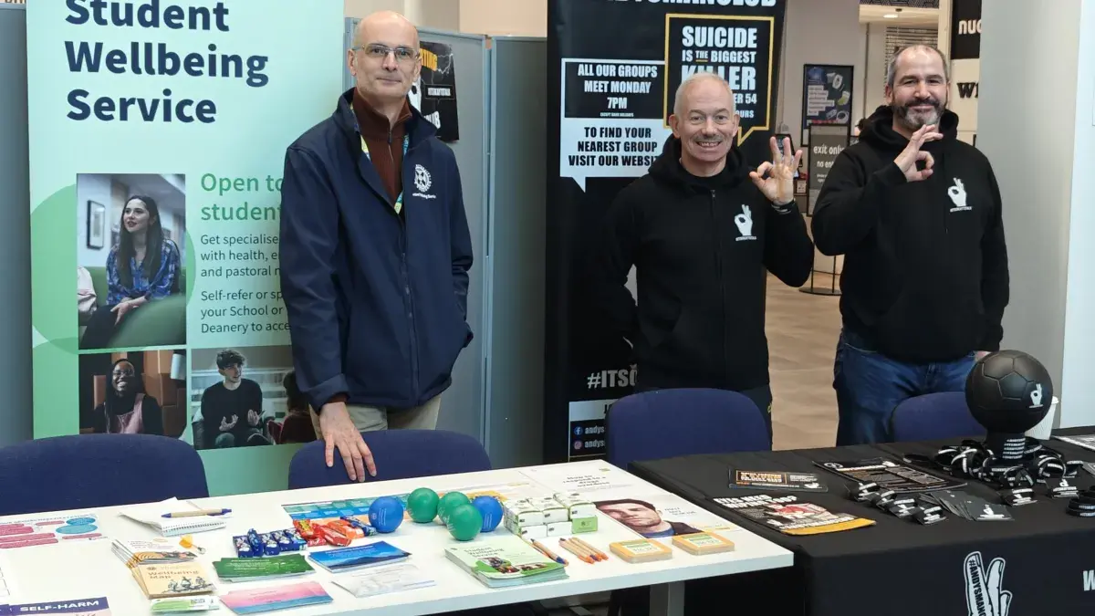 Three smiling men standing behind 2 long tables with literature on the tables and two of the men making the 'OK' sign with their fingers