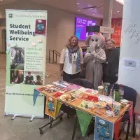 Woman and person dressed in an elephant costume standing next to a banner for the Student Wellbeing Service and behind a rectangular table with branded merchandise on it