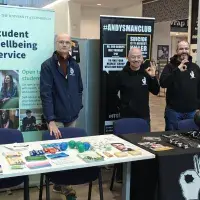 Three smiling men standing behind 2 long tables with literature on the tables and two of the men making the 'OK' sign with their fingers
