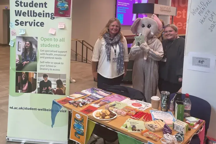 Woman and person dressed in an elephant costume standing next to a banner for the Student Wellbeing Service and behind a rectangular table with branded merchandise on it