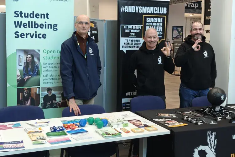 Three smiling men standing behind 2 long tables with literature on the tables and two of the men making the 'OK' sign with their fingers