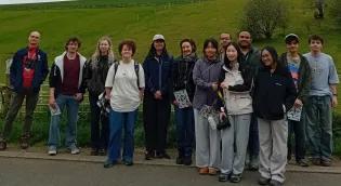 Group photo of 12 students and Chris, wellbeing adviser, standing in a line in front of grassy hill 