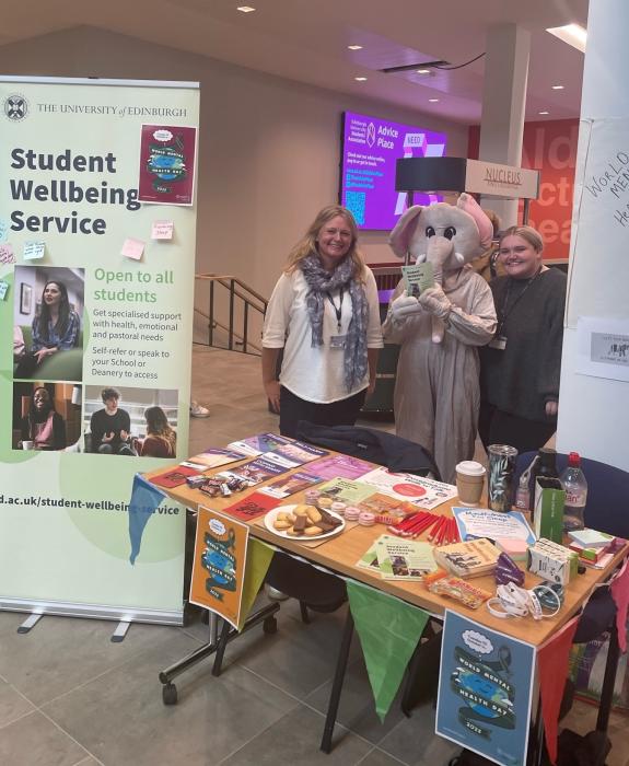 Three people, including one dressed in an elephant costume, standing next to a banner that shows details for the Student Wellbeing Service and a table with informational leaflets on it.